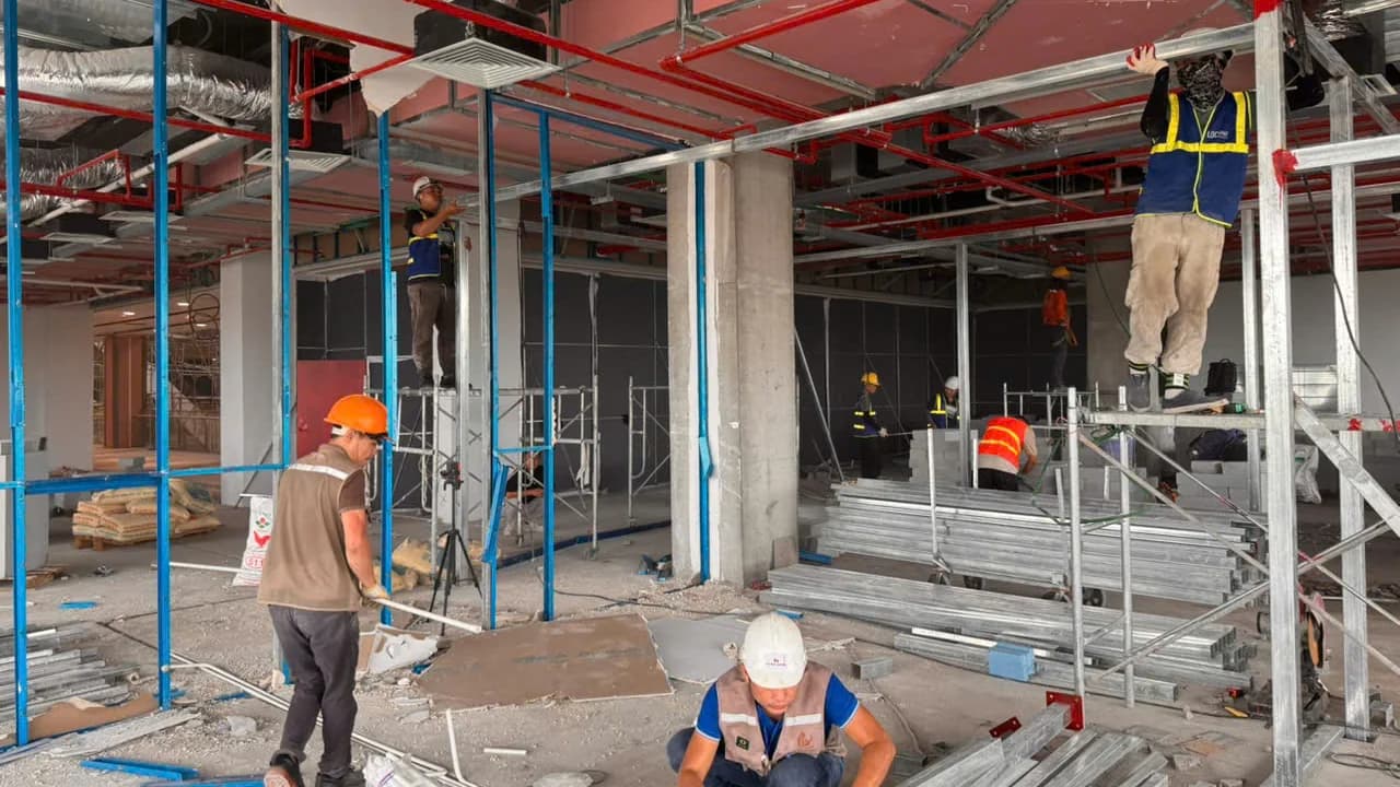 Công trường trong trung tâm thương mại Vincom Đan Phượng: giàn giáo dựng kín mặt bằng, một thợ đứng trên giàn thi công trần, một thợ ngồi lắp khung dưới sàn, phía sau còn nhiều thợ khác đang làm